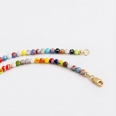 A close-up view of a segment of a multi-colored Millefiori glass bead necklace, lying horizontally on a white surface, finished with a small gold lobster clasp.

