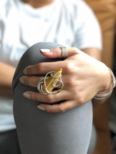 Close-up of a hand wearing a silver ring with a yellow stone on a gray surface.