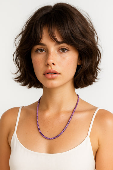 Woman wearing a white tank top and a purple beaded necklace against a white background