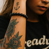 Person with tattoos on arm wearing a black beaded bracelet, with a blurred background.