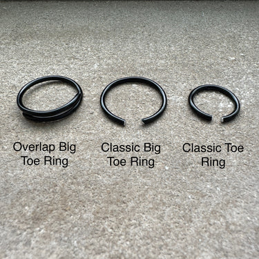 Three black toe rings labeled 'Overlap Big Toe Ring', 'Classic Big Toe Ring', and 'Classic Toe Ring' on a concrete surface. Black anodized aluminum toe ring displayed on stone surface — from the COLORS Collection.