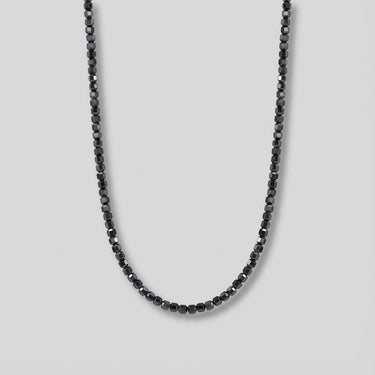 Partial view of Men's necklace featuring small faceted Black Onyx cube beads with polished geometric surfaces, arranged in a curved shape against a light gray background.