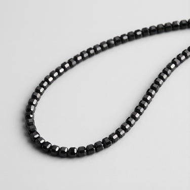 Close-up of a men's necklace featuring small faceted Black Onyx cube beads with polished geometric surfaces, arranged in a curved shape against a light gray background.