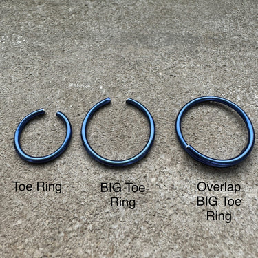 Three blue toe rings labeled 'Toe Ring', 'BIG Toe Ring', and 'Overlap BIG Toe Ring' on a concrete surface. Dark blue anodized aluminum toe rings displayed on stone surface — lightweight, water-friendly design from the COLORS Collection.