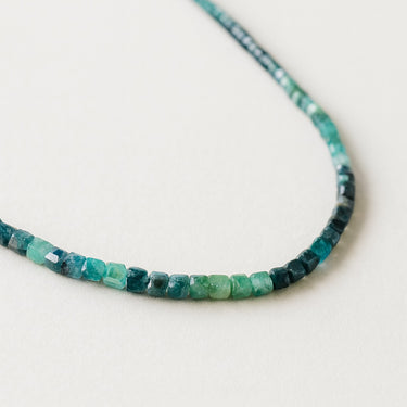 A macro close-up of a tourmaline bead strand showing the transition of natural green, mint, and forest tones in faceted cube shapes.
