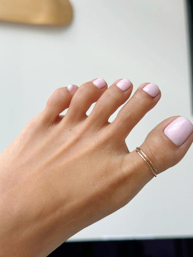 Close-up of a foot wearing a gold big toe overlap ring with pink nail polish on a neutral background