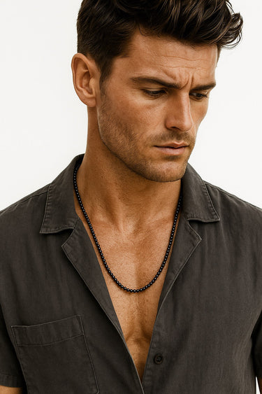 A male model wearing a grey button-down shirt showcasing a sleek strand of 4mm black onyx beads.
