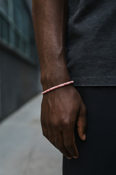 A close-up of a male model's wrist showcasing the muted pink tones and low-profile fit of a 4mm Rhodonite heishi bead bracelet.
