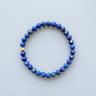 A 6mm deep blue Lapis Lazuli round bead bracelet featuring a single 14K gold-filled accent bead, displayed against a light grey background.
