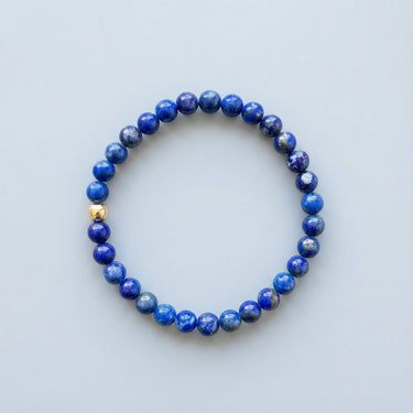 A 6mm deep blue Lapis Lazuli round bead bracelet featuring a single 14K gold-filled accent bead, displayed against a light grey background.
