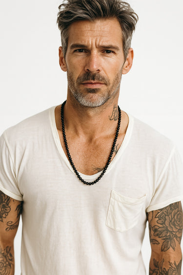 A male model wearing a white v-neck t-shirt showcasing a high-shine 6mm black onyx beaded necklace.
