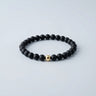 A 6mm matte black onyx round bead bracelet featuring a single 14K gold-filled accent bead, displayed against a light grey background.
