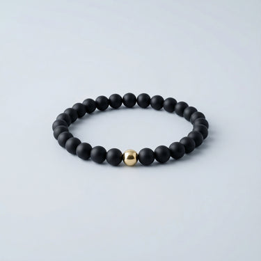 A 6mm matte black onyx round bead bracelet featuring a single 14K gold-filled accent bead, displayed against a light grey background.

