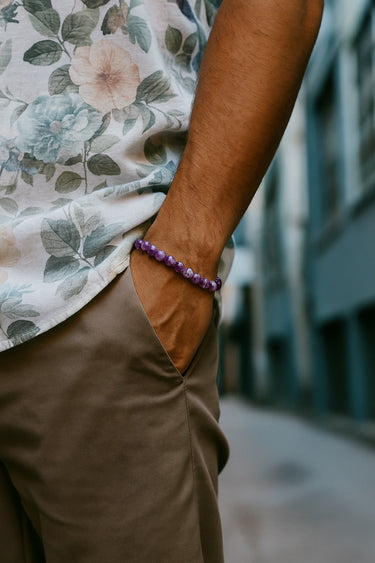 A close-up of a male model's wrist showcasing the bold 8mm scale and rich violet hues of an Amethyst round bead bracelet paired with khaki trousers.
