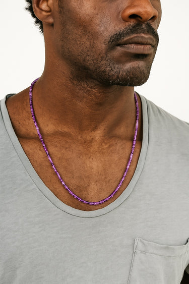 A male model wearing a light grey v-neck t-shirt showcasing the rich purple tones and textured drape of a 4mm amethyst heishi necklace.
