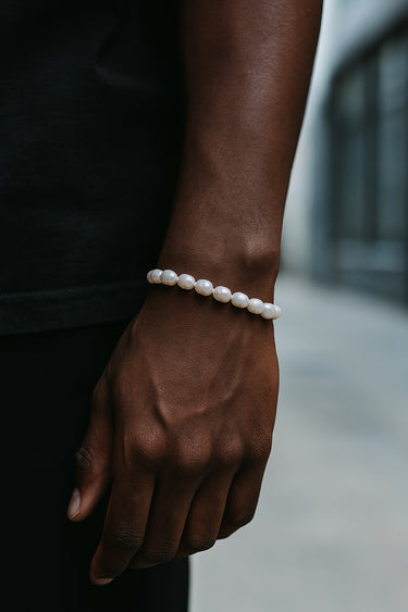 A close-up of a male model's wrist highlighting the unique, organic silhouettes of white baroque pearls against a dark background.
