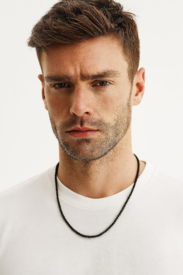 A male model wearing a white t-shirt showcasing the textured, matte finish of a 4mm black lava beaded necklace.
