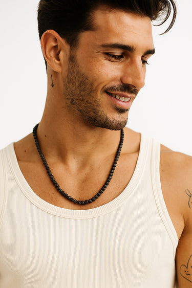 A man in a white ribbed tank top showcasing the drape of a matte black onyx beaded necklace.
