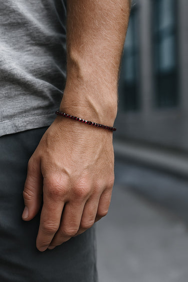 A close-up of a male model's wrist showcasing the sharp geometric silhouette and dark red hues of a 3mm Garnet cube bracelet.
