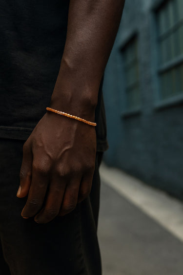 A close-up of a male model's wrist showcasing the delicate geometric silhouette and warm golden glow of a 2mm Sunstone cube bracelet.
