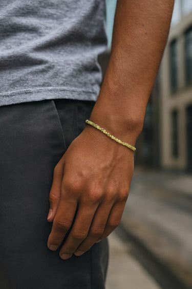 A close-up of a male model's wrist showcasing the delicate geometric silhouette and bright green hues of a 2mm Peridot cube bracelet.
