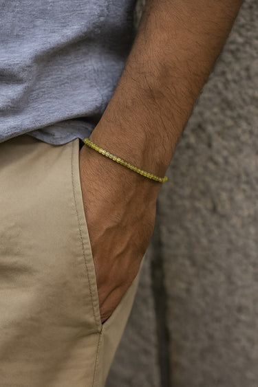 A male model's hand in his pocket, highlighting the low-profile fit and vibrant green color of a 2mm faceted peridot bracelet.

