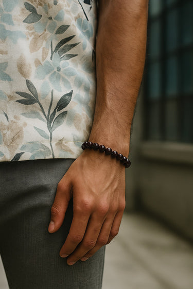 A close-up of a male model's wrist showcasing the bold 8mm scale and rich dark red hues of a Garnet round bead bracelet paired with a patterned floral shirt.
