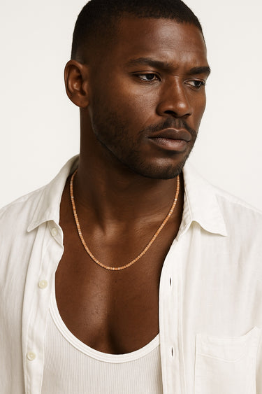 A Black male model wearing an open white button-down shirt showcasing a delicate strand of golden orange faceted sunstone cube beads.
