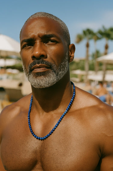 A male model outdoors in a beach setting wearing a single strand of 6mm blue Lapis Lazuli beads.
