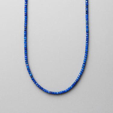 A deep blue faceted Lapis Lazuli cube bead necklace displayed in a U-shape against a white background.
