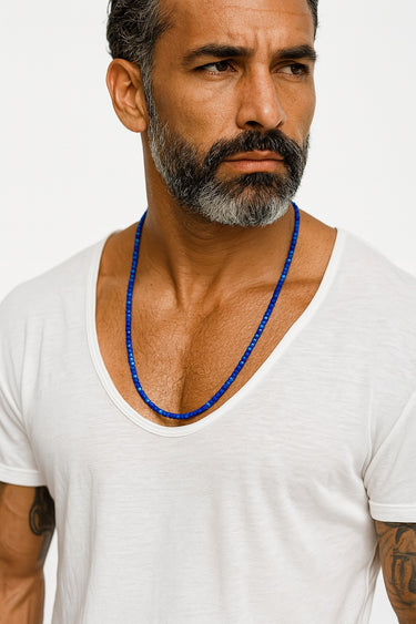 A male model in a white shirt wearing a single strand of small blue faceted Lapis Lazuli cube beads.

