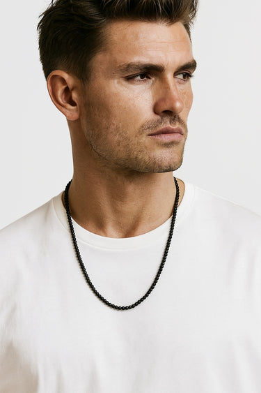 A close-up portrait of a male model in a white t-shirt wearing a single strand of 4mm matte black gemstone beads.
