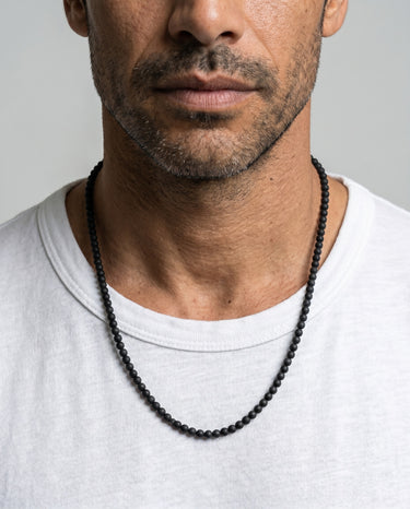 A close-up portrait of a male model in a white t-shirt wearing a single strand of 4mm matte black round gemstone beads.