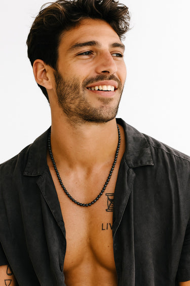 A smiling man in a black open-collar button-down shirt wearing a single strand of matte black onyx beads.
