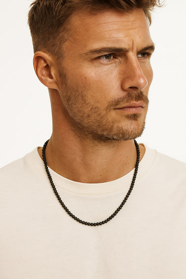 A male model wearing a white t-shirt showcasing the understated, non-reflective finish of a 4mm matte black onyx beaded necklace.

