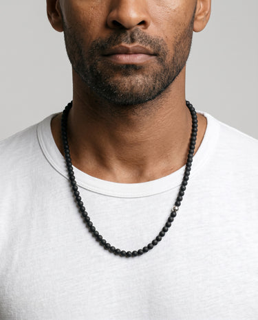 Men’s 6mm matte black onyx bead necklace with an off-centered sterling silver accent bead, worn with a light gray crew neck shirt.