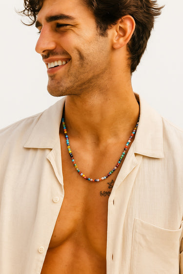 A close-up view of a smiling male model wearing an unbuttoned cream shirt, showcasing the multi-colored Millefiori bead necklace against his skin.
