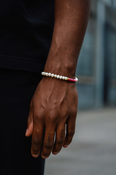 A close-up of a male model's wrist showcasing the contrast between white pearls and pink rhodonite beads against a dark background.
