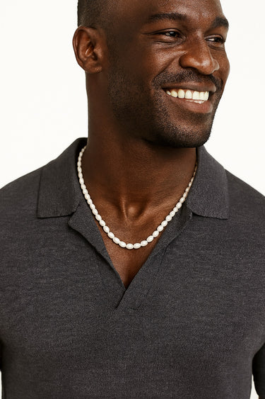 A close-up portrait of a smiling man in a dark polo shirt wearing a strand of medium-sized white freshwater pearls.
