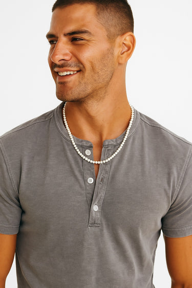 A male model in a grey henley shirt wearing a single strand of small white freshwater pearls.
