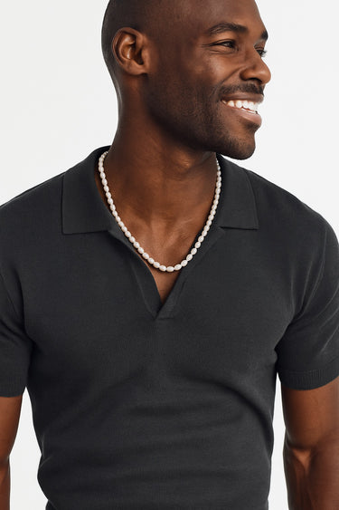 A Black male model wearing a dark charcoal polo shirt showcasing the luster of a freshwater pearl necklace.
