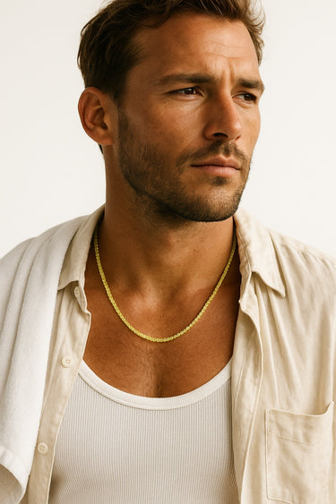 A male model wearing a cream-colored button-down shirt over a white tank top, showcasing a delicate strand of lime-green faceted peridot cube beads.
