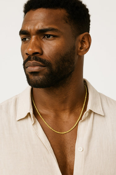 A portrait of a Black male model in a cream shirt, featuring a single strand of small, faceted green peridot beads around the neck.
