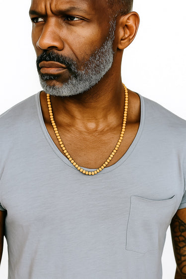 A close-up of a man in a light grey t-shirt showcasing the warm, matte tones of a golden sandalwood necklace.
