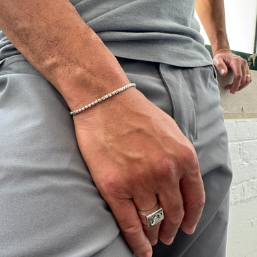 A lifestyle shot of a male model wearing a 2.5mm sterling silver cube chain bracelet, highlighting its slim, geometric profile paired with grey trousers.
