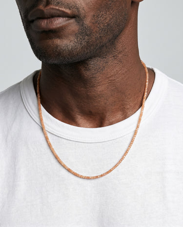 A male model wearing an open white t-shirt showcasing a delicate strand of golden orange faceted sunstone cube beads.