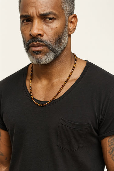 A male model wearing a white t-shirt showcasing the warm, golden luster of a 6mm Tiger's Eye beaded necklace.
