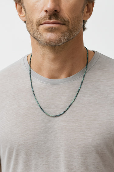 A male model wearing a grey crewneck t-shirt showcasing a single strand of small faceted tourmaline cube beads.
