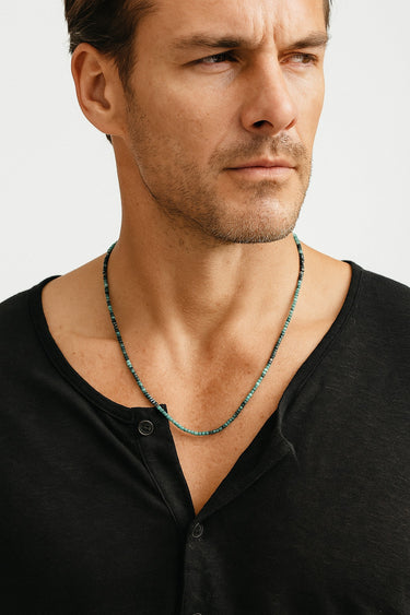 A male model in a black henley shirt showcasing the deep green tones of a faceted tourmaline cube necklace.

