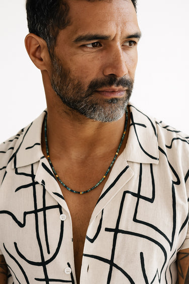 A close-up portrait of a bearded male model wearing a patterned button-down shirt and a strand of faceted tourmaline beads with gold accents.

